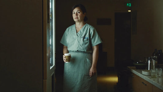 A nurse stands in a dim hospital break room at 3 AM, tired but composed, holding a coffee cup