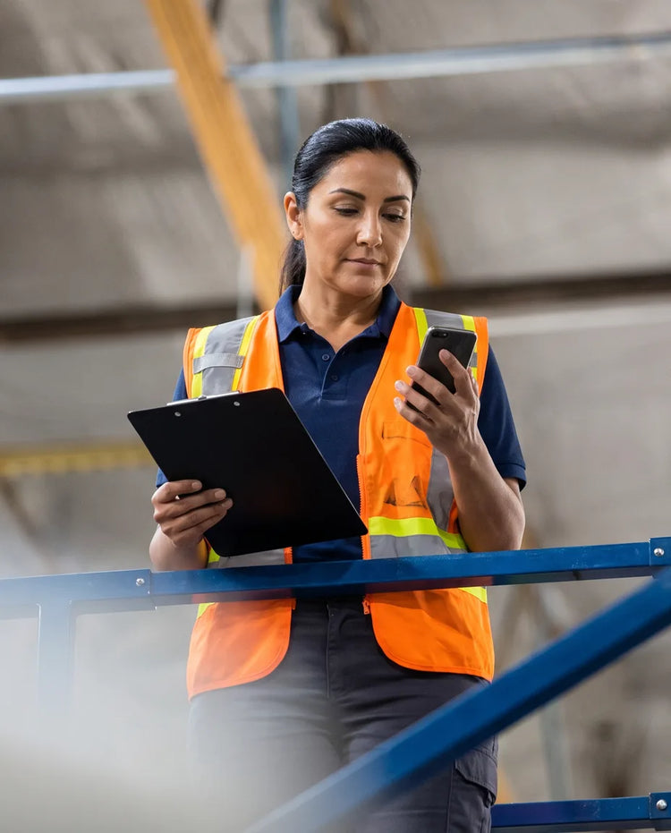 A warehouse supervisor in a hi-vis vest checking a clipboard and phone on a mezzanine walkway