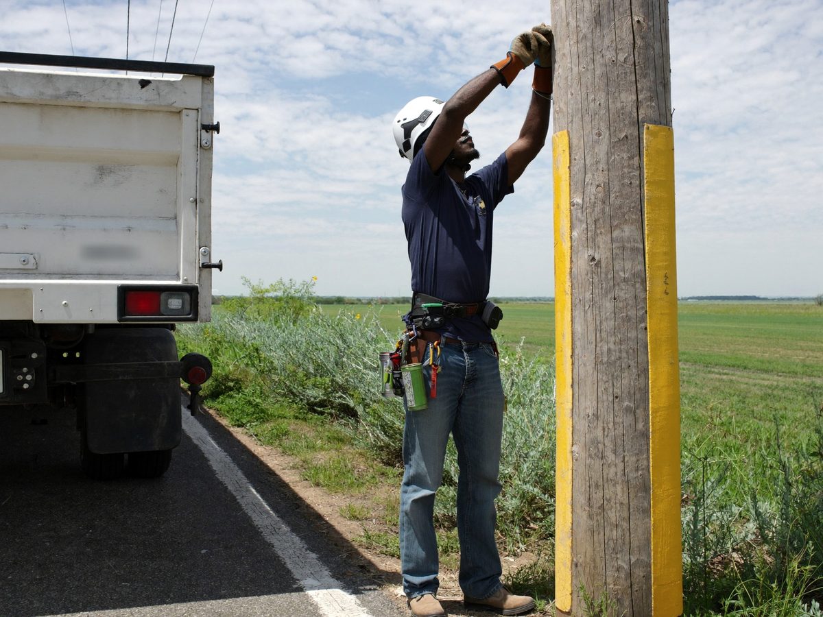 A lineman beside his bucket truck on a country road, reaching up a wooden utility pole