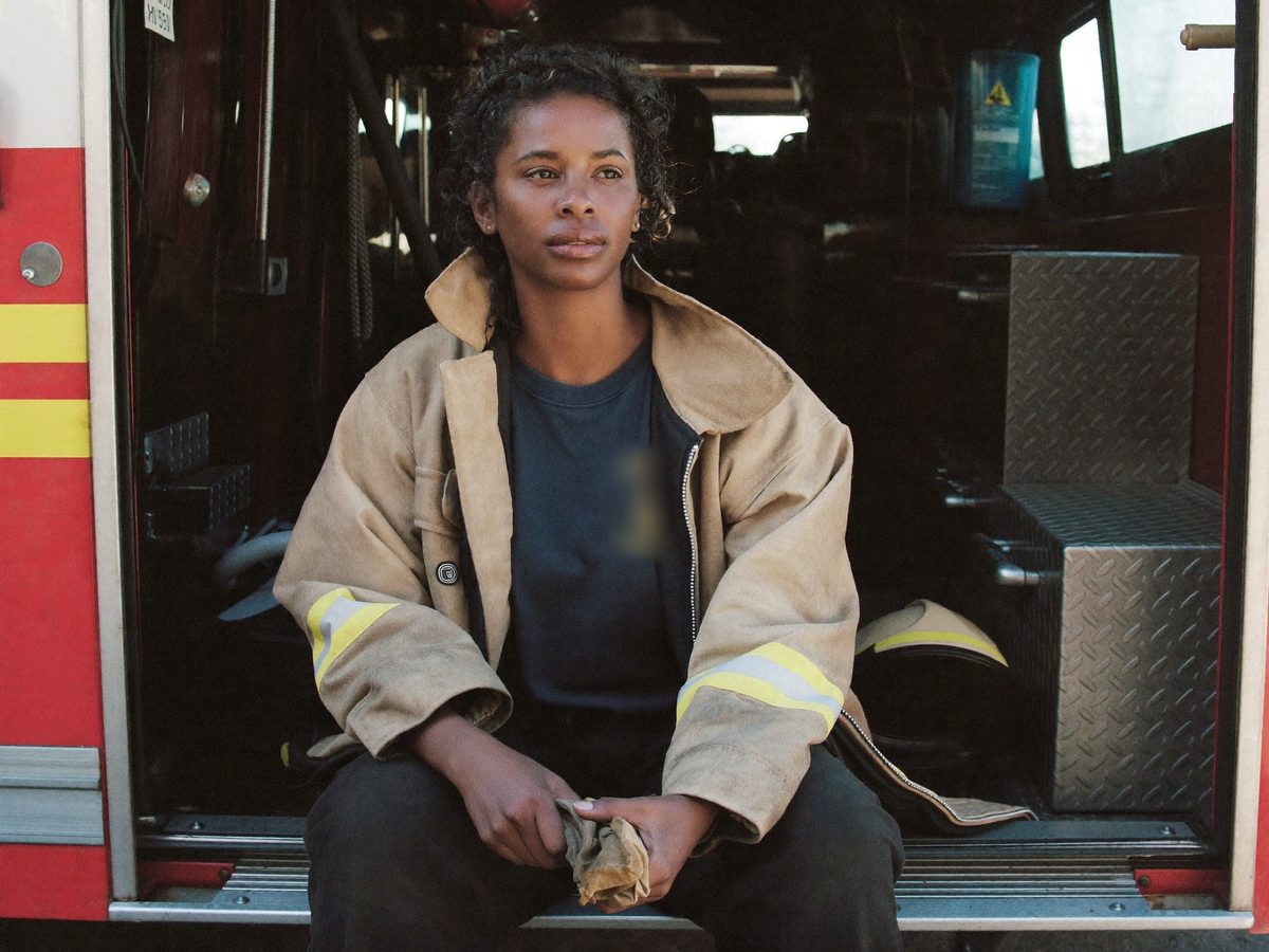 A firefighter in her turnout coat sitting on the rear step of the engine after a call