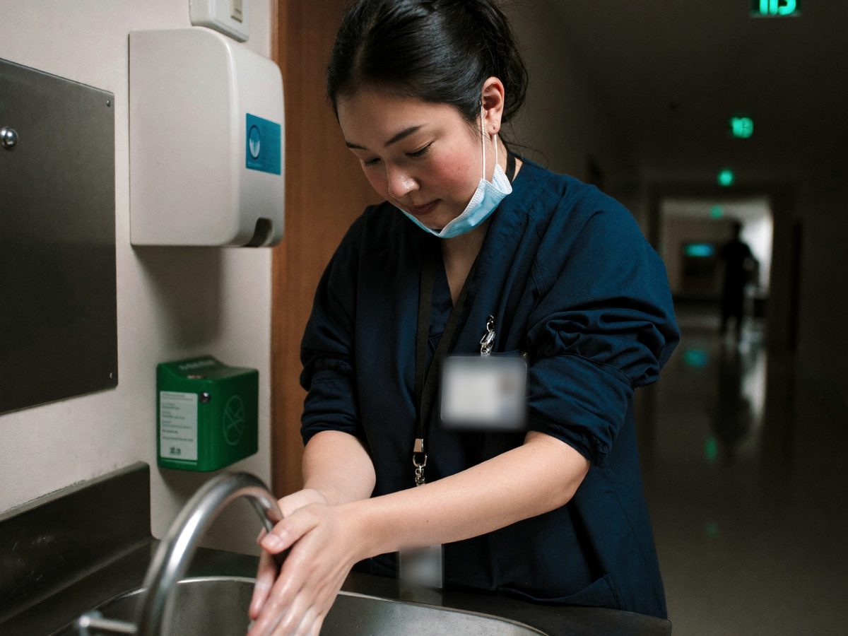 A nurse in navy scrubs washing her hands at a hospital corridor sink